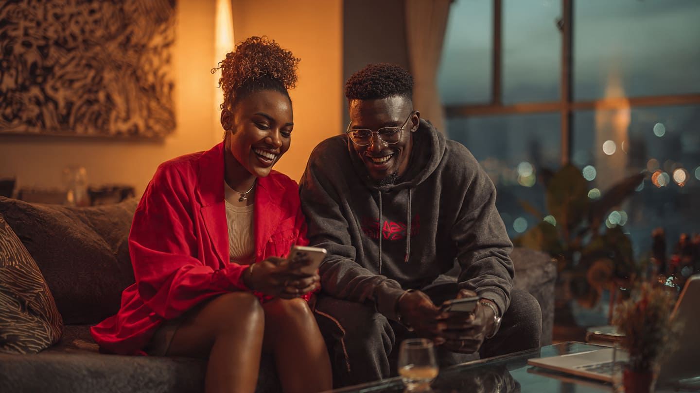Two young adults smiling and using smartphones together on a couch in a warmly lit modern apartment.