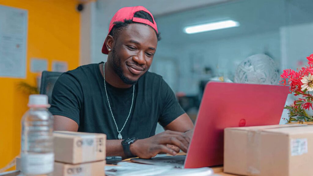 A Nigerian entrepreneur packing orders in a bright office with a laptop showing a successful way to convert Instagram comments to sales funnel