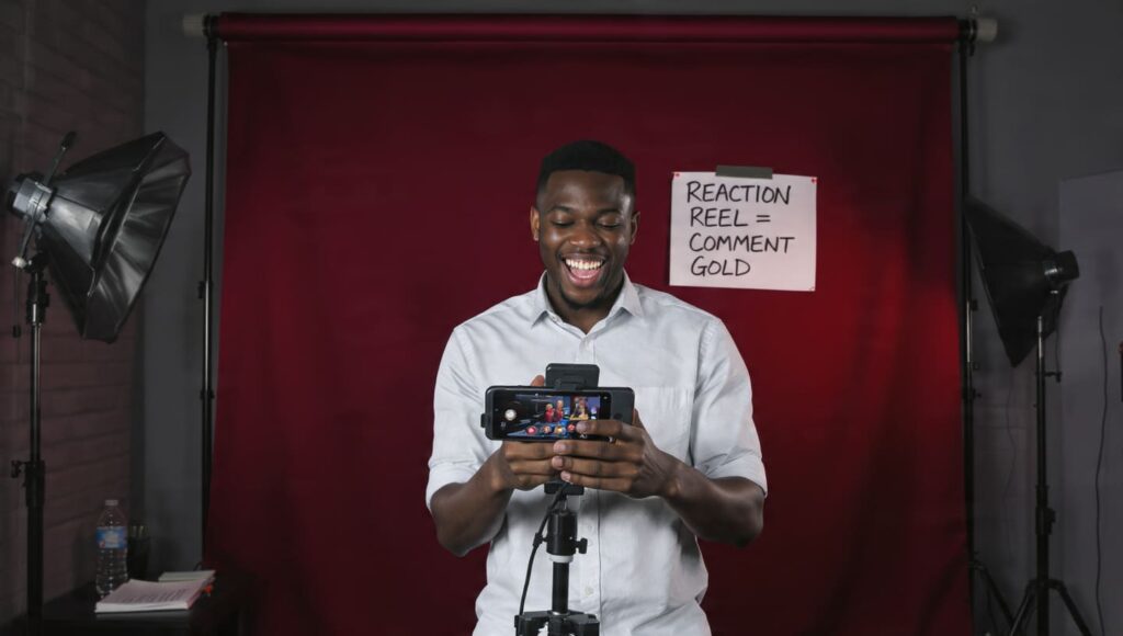 A content creator laughing while filming a reaction video on his smartphone in front of a professional red backdrop.